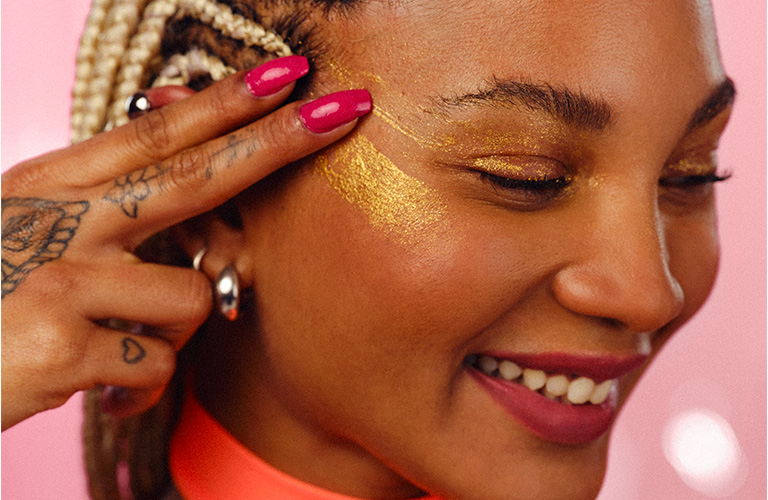 foto de uma mulher sorrindo e passando o Gel Glitter Faces nas têmporas do rosto com os dedos indicador e médio.