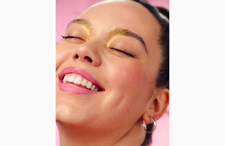 foto de uma mulher sorrindo para câmera, de olhos fechados, mostrando sua maquiagem de carnaval com o Gel Glitter Faces aplicado no canto interno e acima das pálpebras fixas dos olhos.