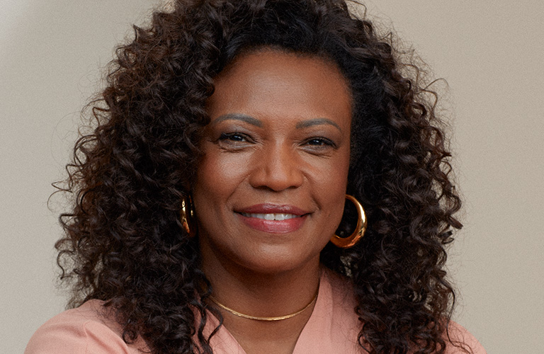 Image of a smiling woman aged 50 to 60 with curly dark brown hair.  She wears a light pink blouse, gold ring and necklace, light eye makeup and a pink lip gloss effect.